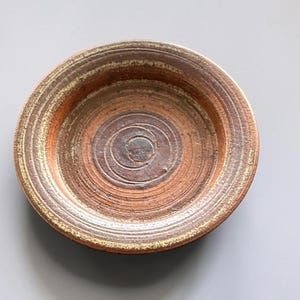 Könnte beinhalten: Eine runde, flache Keramikschale mit einer terrakottafarbenen Basis und konzentrischen kreisförmigen Rillen. Die Oberfläche weist ein strukturiertes, erdiges Finish mit variierenden braunen und gedämpften gelben Tönen auf.
