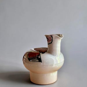 Peut inclure: Un pichet en céramique avec une finition blanc cassé brillante et une base cylindrique. Le corps présente une forme arrondie asymétrique avec un bec court décentré. Des motifs abstraits peints à la main en rouge et noir ornent le dessus.