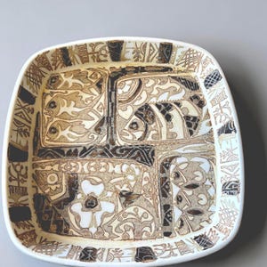 May include: A square ceramic dish with rounded corners, featuring a cream-coloured base decorated with intricate brown and black line art. The design includes stylised fish, floral motifs, and geometric patterns arranged in a segmented grid layout.