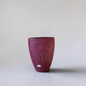 May include: A matte dark magenta vase with a tapered base and a wide, oval-shaped opening. The exterior features a raised, embossed design of two ginkgo leaves on the front surface. A small, circular white sticker is attached to the lower front.