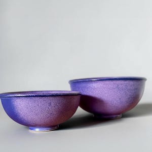 May include: Two ceramic bowls with a rounded shape and a footed base. The bowls feature a vibrant purple and blue gradient glaze with a speckled texture. The interior and exterior surfaces show a smooth finish with a dark blue rim along the top edge.