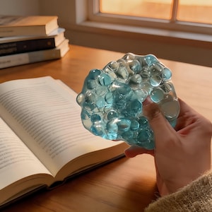 May include: A hand holds a translucent, blue-coloured, amorphous object with a bubbly texture. An open book and other books are in the background. The object appears to be made of many small, spherical shapes. The lighting suggests a window.