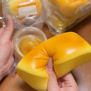 May include: A close-up of a yellow and orange squishy toy being squeezed. The toy is shaped like a loaf of bread. Other similar toys are visible in the background, some in clear plastic packaging.