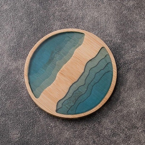 Puede incluir: Posavasos redondo de madera con un diseño en capas en tonos azules y turquesas que recuerda a un paisaje costero. El posavasos tiene un borde de madera natural y un diseño central con diferentes tonos azules y turquesas, creando profundidad visual.