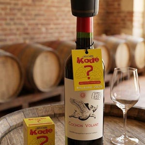 Peut inclure: Une bouteille de vin rouge avec un bouchon de sécurité noir. L'étiquette indique "Cochon Volant" avec une illustration de cochon ailé. Une étiquette jaune avec "Bouchon Kodé" et un point d'interrogation est accrochée à la bouteille. Un verre à vin est à proximité.