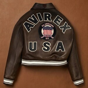 Op de afbeelding: Bruine leren jas met witte en zwarte letters en patches. De jas heeft het woord "AVIREX" over de schouders, een patch met de Amerikaanse vlag en het woord "USA". De jas heeft wit en zwart gestreepte manchetten.