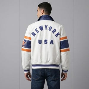 K&ouml;nnte beinhalten: Cremefarbene Lederjacke mit blauen und orangefarbenen Akzenten. Auf dem R&uuml;cken steht "NEW YORK USA" in blauen Buchstaben. Die Jacke hat einen blauen Kragen und gestreifte B&uuml;ndchen und Saum.
