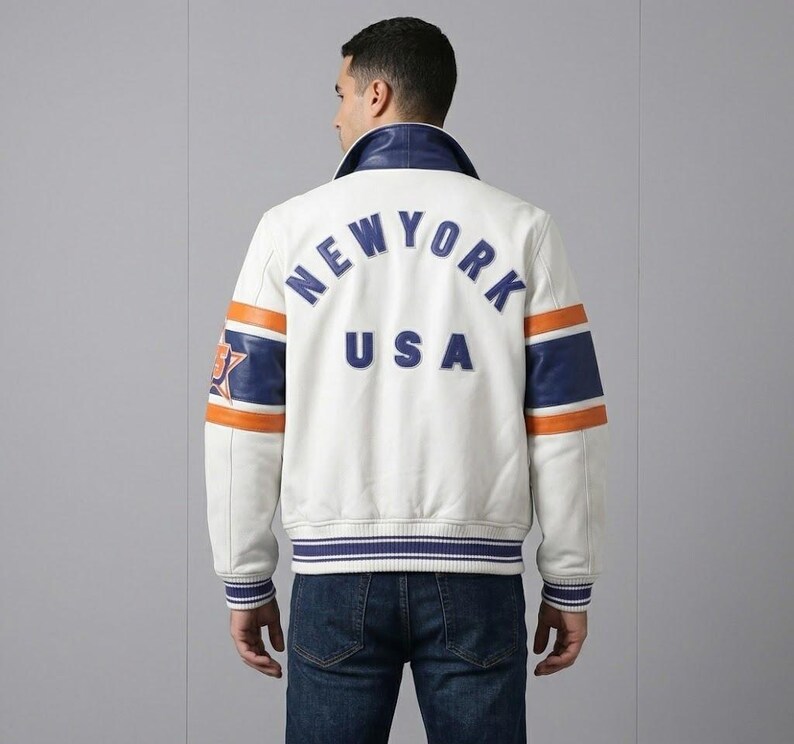 K&ouml;nnte beinhalten: Cremefarbene Lederjacke mit blauen und orangefarbenen Akzenten. Auf dem R&uuml;cken steht "NEW YORK USA" in blauen Buchstaben. Die Jacke hat einen blauen Kragen und gestreifte B&uuml;ndchen und Saum.