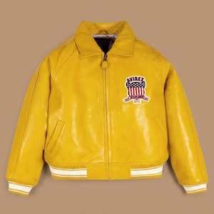 May include: A yellow leather jacket with a zip-up front, a collar, and a patch on the chest. The patch features a shield design with red, white, and blue colors. The jacket has white stripes on the cuffs and hem.