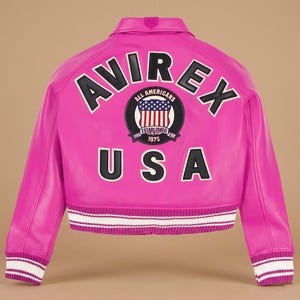 Op de afbeelding: Een felroze leren jas met het woord "AVIREX" in witte letters met zwarte contouren op de rug. Daaronder een cirkelvormige patch met "ALL AMERICANS" en "ESTABLISHED 1975". Het woord "USA" staat eronder. De manchetten zijn wit en roze gestreept.