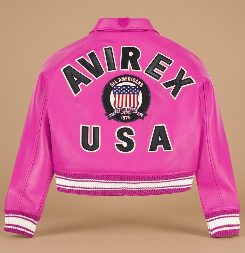 Op de afbeelding: Een felroze leren jas met het woord "AVIREX" in witte letters met zwarte contouren op de rug. Daaronder een cirkelvormige patch met "ALL AMERICANS" en "ESTABLISHED 1975". Het woord "USA" staat eronder. De manchetten zijn wit en roze gestreept.