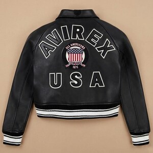 Op de afbeelding: Zwarte leren jas met witte letters "AVIREX" en "USA". Een cirkelvormige patch toont de Amerikaanse vlag en de woorden "ALL AMERICANS ESTABLISHED 1975". De jas heeft wit en zwart gestreepte manchetten en zoom.
