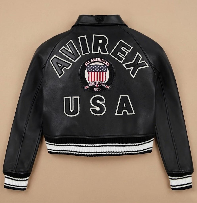 Op de afbeelding: Zwarte leren jas met witte letters "AVIREX" en "USA". Een cirkelvormige patch toont de Amerikaanse vlag en de woorden "ALL AMERICANS ESTABLISHED 1975". De jas heeft wit en zwart gestreepte manchetten en zoom.