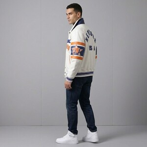 K&ouml;nnte beinhalten: Cremefarbene Collegejacke mit blauen und orangefarbenen Akzenten. Die Jacke hat ein Sternemblem und den Text "NEW YORK USA". Sie hat einen blauen Kragen und Manschetten sowie einen gestreiften Saum. Die Person tr&auml;gt dunkelblaue Jeans und wei&szlig;e Turnschuhe.