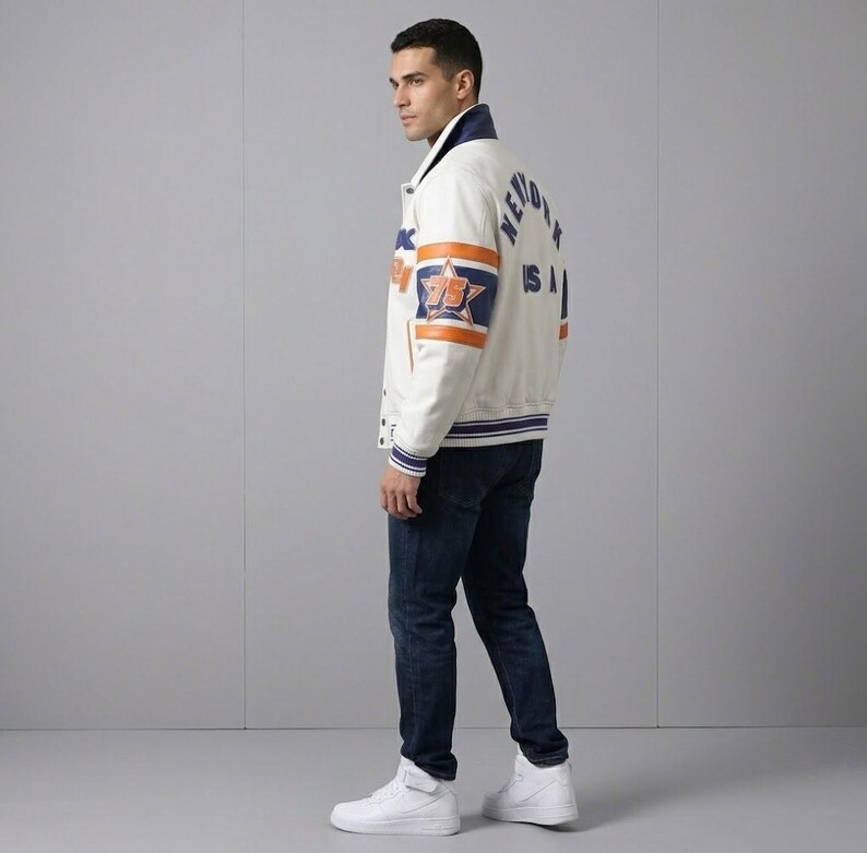 K&ouml;nnte beinhalten: Cremefarbene Collegejacke mit blauen und orangefarbenen Akzenten. Die Jacke hat ein Sternemblem und den Text "NEW YORK USA". Sie hat einen blauen Kragen und Manschetten sowie einen gestreiften Saum. Die Person tr&auml;gt dunkelblaue Jeans und wei&szlig;e Turnschuhe.