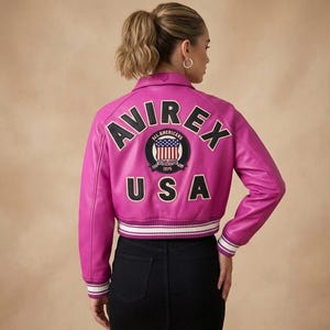 Op de afbeelding: Een felroze leren jas met het woord "AVIREX" over de rug, boven een cirkelvormige patch met "ALL AMERICANS" en "USA" eronder. De jas heeft een wit en zwart gestreepte zoom en manchetten.