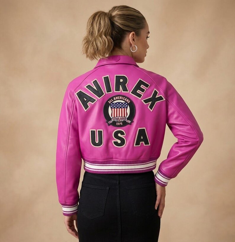 Op de afbeelding: Een felroze leren jas met het woord "AVIREX" over de rug, boven een cirkelvormige patch met "ALL AMERICANS" en "USA" eronder. De jas heeft een wit en zwart gestreepte zoom en manchetten.