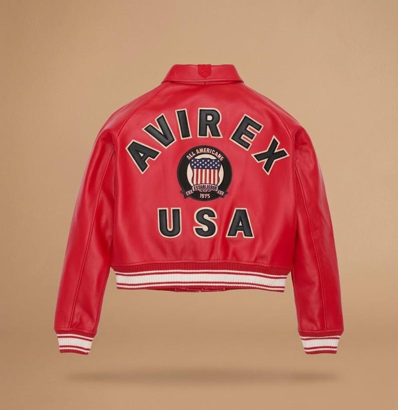 Op de afbeelding: Rode leren jas met het woord "AVIREX" in zwarte letters op de rug. Daaronder een cirkelvormige patch met "ALL AMERICANS" en "USA". De jas heeft een wit en rood gestreepte zoom. Klassieke bomberstijl.