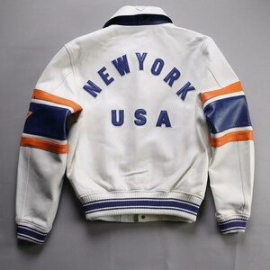 K&ouml;nnte beinhalten: Wei&szlig;e Lederjacke mit blauen und orangefarbenen Akzenten. Auf dem R&uuml;cken steht "NEW YORK USA" in Blau. Die &Auml;rmel haben blaue und orangefarbene Streifen. Gerippter Kragen und B&uuml;ndchen. Klassischer Bomberstil.