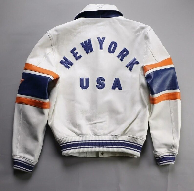 K&ouml;nnte beinhalten: Wei&szlig;e Lederjacke mit blauen und orangefarbenen Akzenten. Auf dem R&uuml;cken steht "NEW YORK USA" in Blau. Die &Auml;rmel haben blaue und orangefarbene Streifen. Gerippter Kragen und B&uuml;ndchen. Klassischer Bomberstil.