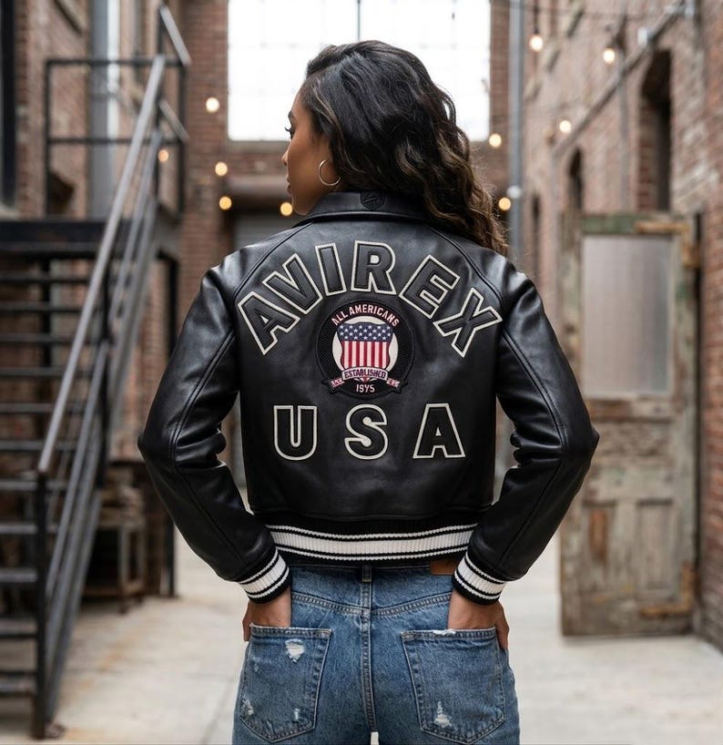 Op de afbeelding: Zwarte leren jas met witte bies en de tekst "AVIREX" en "USA" in het wit. De jas heeft een patch met een rood-wit-blauw ontwerp. De jas wordt gedragen met een blauwe spijkerbroek.