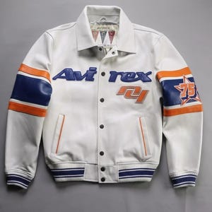 K&ouml;nnte beinhalten: Wei&szlig;e Lederjacke mit blauen und orangefarbenen Akzenten. Sie zeigt den Markennamen "Avirex" in Blau und die Zahl "75" in einem Stern auf dem &Auml;rmel. Die Jacke hat eine Knopfleiste und gestreifte B&uuml;ndchen.