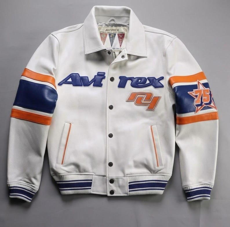 K&ouml;nnte beinhalten: Wei&szlig;e Lederjacke mit blauen und orangefarbenen Akzenten. Sie zeigt den Markennamen "Avirex" in Blau und die Zahl "75" in einem Stern auf dem &Auml;rmel. Die Jacke hat eine Knopfleiste und gestreifte B&uuml;ndchen.