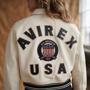 Op de afbeelding: Cr&egrave;mekleurige bomberjack met zwarte letters die "AVIREX" en "USA" spellen. Een cirkelvormige patch toont de woorden "ALL AMERICAN" en een schildontwerp met sterren en strepen. De jas heeft een zwart-witte gestreepte zoom.