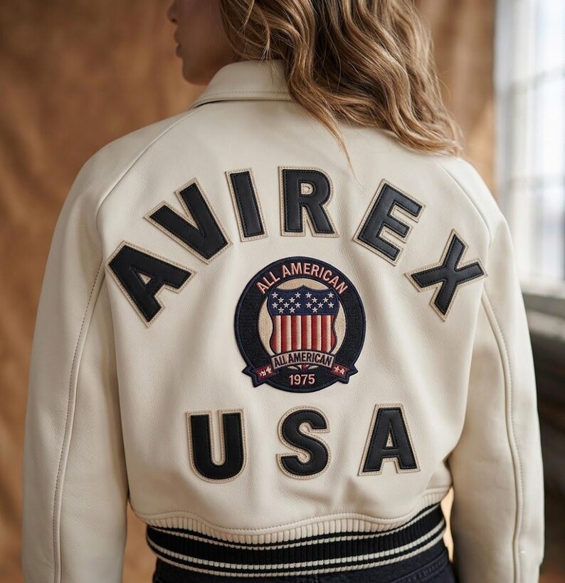 Op de afbeelding: Cr&egrave;mekleurige bomberjack met zwarte letters die "AVIREX" en "USA" spellen. Een cirkelvormige patch toont de woorden "ALL AMERICAN" en een schildontwerp met sterren en strepen. De jas heeft een zwart-witte gestreepte zoom.