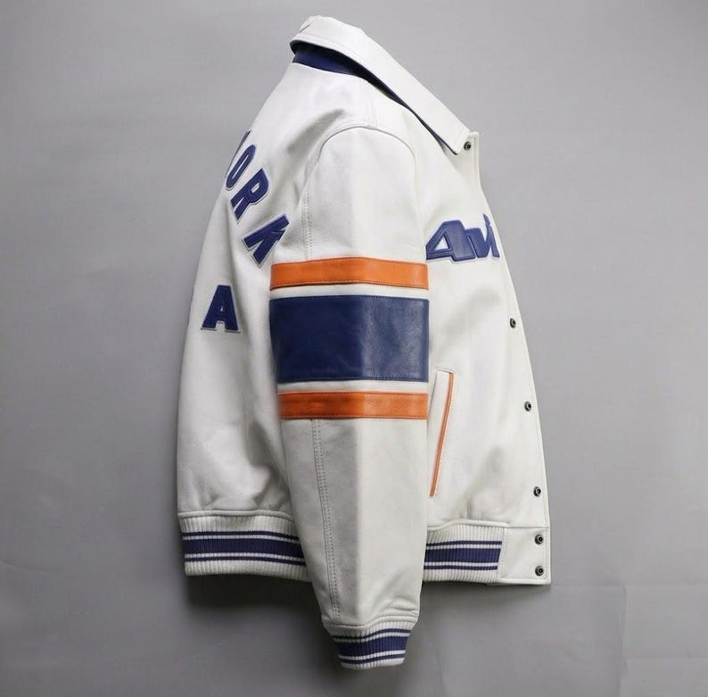 K&ouml;nnte beinhalten: Wei&szlig;e Lederjacke mit blauen und orangefarbenen Akzenten. Auf der Jacke steht in Blau "NEW YORK" auf dem R&uuml;cken und "AM" auf der Vorderseite. Die &Auml;rmel haben ein blau-oranges Streifendesign. Sie hat eine Knopfleiste und einen gerippten Kragen und B&uuml;ndchen.