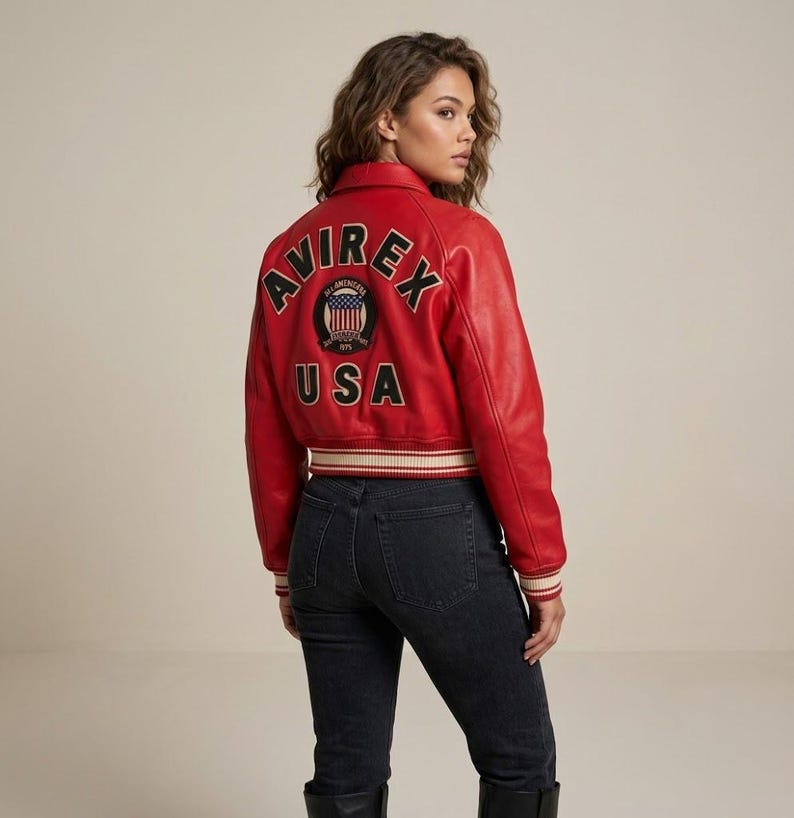 Op de afbeelding: Rood leren jack met het woord "AVIREX" op de rug, boven een cirkelvormig embleem en het woord "USA". De jas heeft een geribbelde zoom en manchetten met witte en beige strepen. Het model draagt een zwarte jeans.