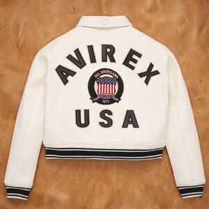 Op de afbeelding: Cr&egrave;mekleurige bomberjack met zwart-witte gestreepte boorden. De achterkant heeft het woord "AVIREX" in zwarte letters boven een cirkelvormig embleem met de Amerikaanse vlag en de tekst "ALL AMERICANS ESTABLISHED 1975 USA".
