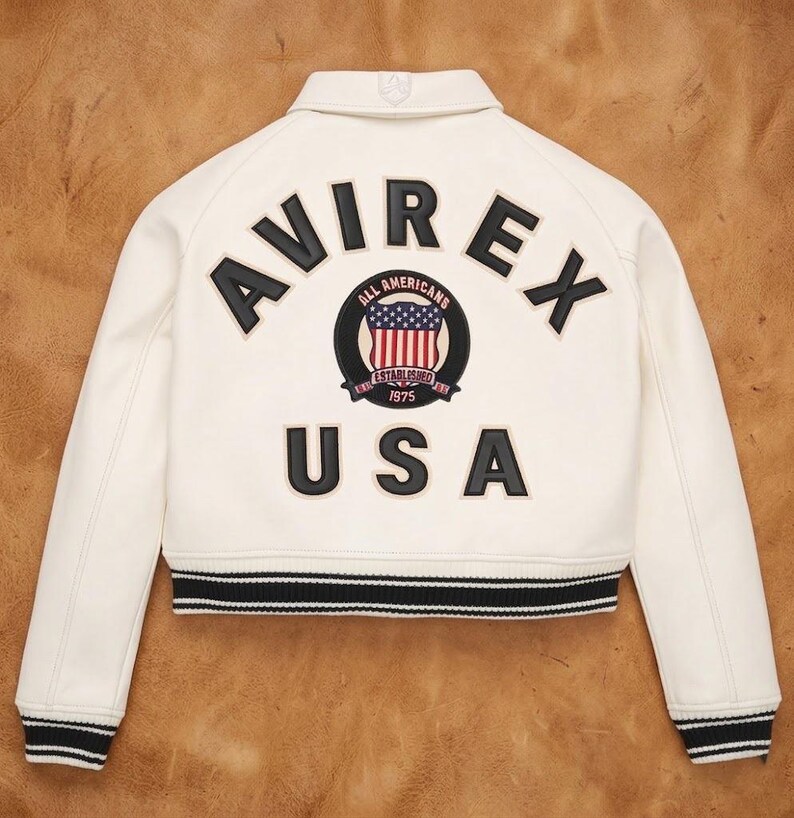 Op de afbeelding: Cr&egrave;mekleurige bomberjack met zwart-witte gestreepte boorden. De achterkant heeft het woord "AVIREX" in zwarte letters boven een cirkelvormig embleem met de Amerikaanse vlag en de tekst "ALL AMERICANS ESTABLISHED 1975 USA".