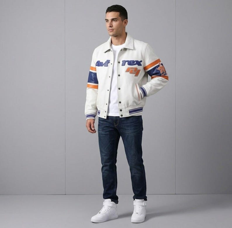K&ouml;nnte beinhalten: Cremefarbene Collegejacke mit blauen und orangefarbenen Akzenten. Die Jacke tr&auml;gt den Text "Avirex" und "74" in blauen und orangefarbenen Buchstaben. Die &Auml;rmel haben blaue und orangefarbene Streifen. Die Person tr&auml;gt ein wei&szlig;es T-Shirt, blaue Jeans und wei&szlig;e Turnschuhe.