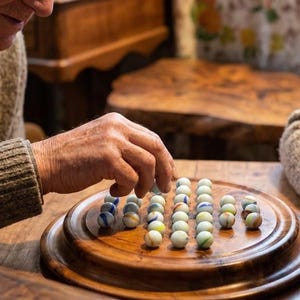 May include: A wooden solitaire board game with marbles. The game is set on a wooden table with a wooden box and a cup of tea. Two people are playing the game.