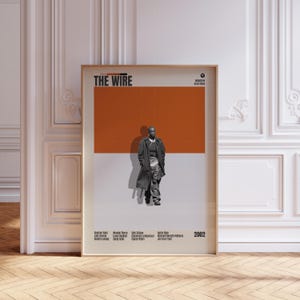 May include: Framed poster featuring the title "THE WIRE" in bold white letters. The poster has a color block design with a man in a long coat standing in front of an orange and gray background. The poster includes the year 2002 and the names of the cast.