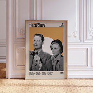 May include: A framed poster for the film "The 39 Steps" in a gold frame. The poster has a black and white image of a man and woman, set against an orange and white backdrop. The title and cast details are displayed in a classic font.