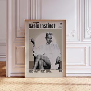 May include: A framed movie poster for Basic Instinct featuring a black and white photograph of a person sitting with legs crossed, wearing a white turtleneck and blazer. The text includes the title, director, cast names, and the year 1992.
