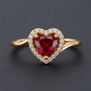 Op de afbeelding: Een gouden ring met een centrale hartvormige rode edelsteen, omringd door een halo van kleine witte, rond geslepen stenen. De ringband is glad en gepolijst en loopt iets taps toe waar deze aansluit op de hartvormige zetting in het midden.