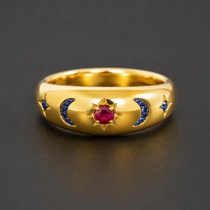 May include: A gold band ring featuring a central star motif set with a round red gemstone. The band is decorated with two crescent moon shapes and two four-pointed star shapes, all inlaid with small blue gemstones surrounding the center.