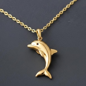 May include: A gold-colored dolphin pendant necklace. The dolphin is sculpted with a smooth finish, featuring a small black eye. The necklace chain is also gold-colored, with a delicate link design. The pendant hangs from a small loop attached to the chain.