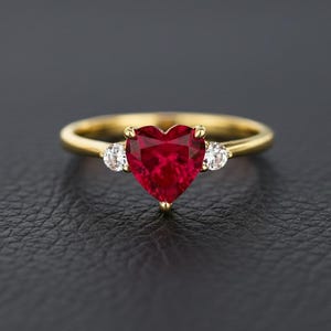 Puede incluir: Un anillo de oro con una piedra preciosa roja oscura en forma de corazón como pieza central. Dos piedras preciosas blancas redondas más pequeñas flanquean la piedra en forma de corazón. El anillo está sobre un fondo oscuro.