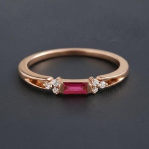 May include: Rose gold ring featuring a rectangular ruby center stone and small diamond accents. The band has a delicate design with cut-out details on each side of the center stone. The ring is set against a dark background.