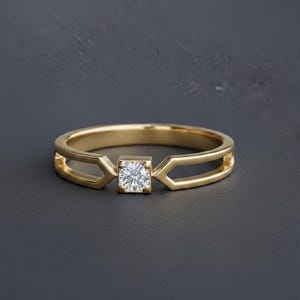May include: A gold ring featuring a single square-set clear gemstone at the centre. The band has a split design on both sides of the stone, creating an open geometric shape that connects to the central setting holding the faceted white stone.