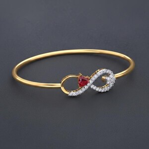 Peut inclure: Un bracelet jonc doré avec un motif de symbole infini. Le symbole infini comporte une pierre précieuse rouge en forme de cœur et est incrusté de petites pierres précieuses claires. Le bracelet est posé sur un fond gris foncé.
