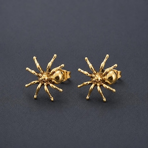 May include: A pair of gold-toned spider-shaped stud earrings. Each earring features a central rounded body with eight radiating legs, showcasing a polished metallic finish and a detailed, textured surface design on the legs and abdomen.