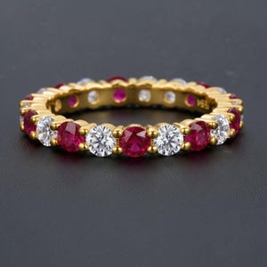 Op de afbeelding: Een gouden ring met afwisselend rode en heldere edelstenen. De ring heeft een doorlopend patroon van rond geslepen stenen gezet in een gepolijste gouden band. De edelstenen zijn gelijkmatig verdeeld.