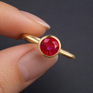 Può includere: Un anello in oro con una pietra preziosa rossa vibrante, rotonda e sfaccettata, montata in una lunetta liscia. La fascia è in oro lucido con un profilo semplice e arrotondato, che mette in risalto la pietra rossa come punto focale.