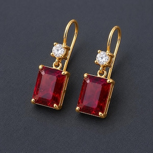 May include: A pair of gold drop earrings featuring a rectangular-cut deep red gemstone suspended below a single round brilliant-cut clear stone. The earrings are attached to gold shepherd hook wires, showcasing a polished metal finish throughout.