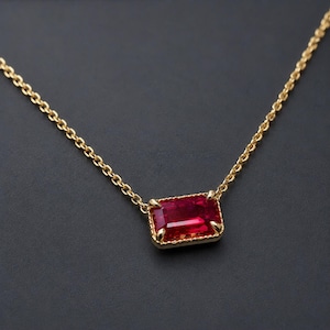 May include: A gold chain necklace featuring a rectangular, emerald-cut red gemstone pendant. The vibrant red stone is set in a gold metal frame with four prongs, suspended from a delicate gold link chain that extends to both sides of the frame.
